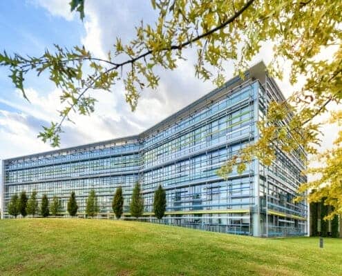 Exterior of a modern angular glass fronted office building in a spring landscape with green lawns and trees under a blue cloudy sky